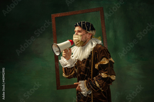 Portrait of medieval young man in vintage clothing, golden face mask shouting with rupor in frame on dark background. Royal person protected from covid. Concept of comparison of eras, modern, fashion. photo