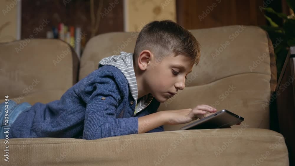 Self-education at home concept where handsome concentrated modern smart boy lying on domestic couch and uses tablet device to look for apps for school Self-education at home concept where handsome concentrated modern smart boy lying on domestic couch and uses tablet device to look for apps for school
