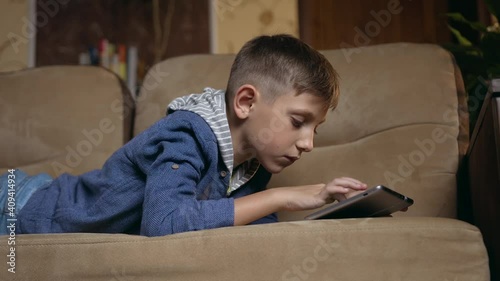 Self-education at home concept where handsome concentrated modern smart boy lying on domestic couch and uses tablet device to look for apps for school photo