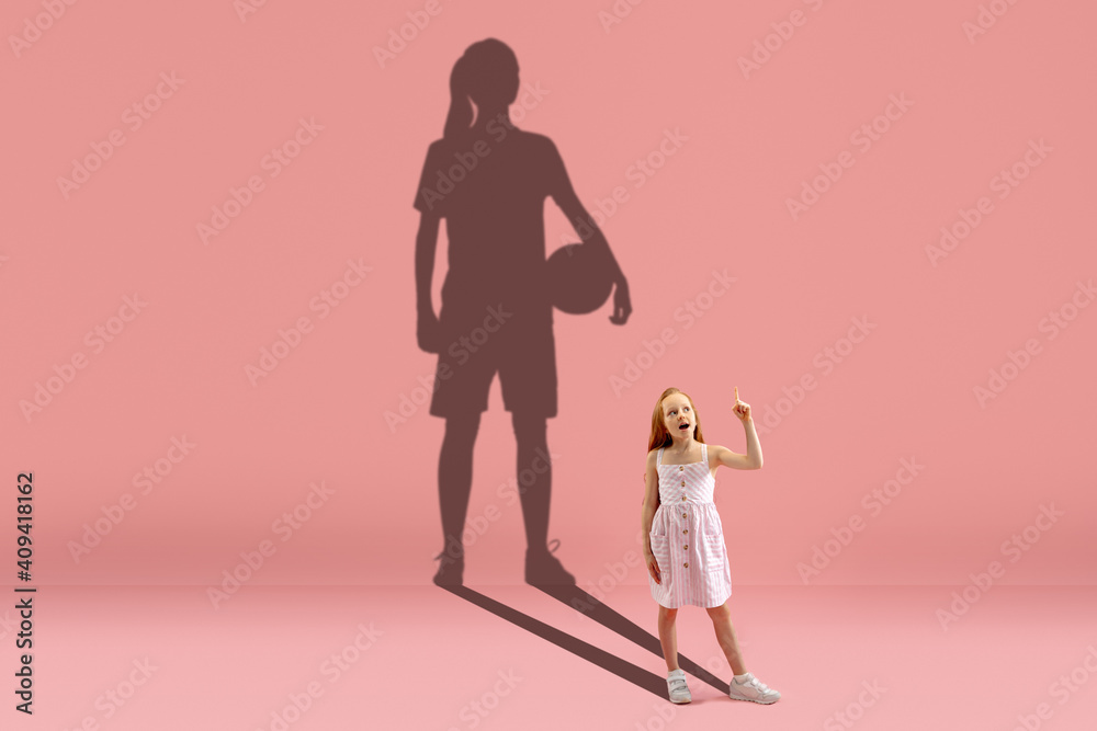 Childhood and dream about big and famous future. Conceptual image with girl and drawned shadow of basketball, soccer female player on coral pink background. Childhood, dreams, education concept. Childhood and dream about big and famous future. Conceptual image with girl and drawned shadow of basketball, soccer female player on coral pink background. Childhood, dreams, education concept.