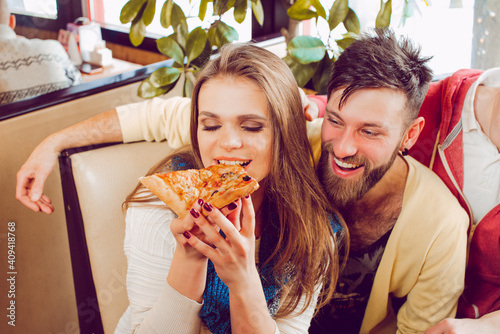 The company of guys in the pizzeria. A guy hugs a girl who bites a slice of pizza. Couple at the pizzeria