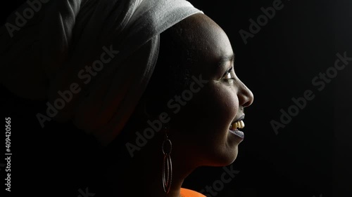 low key portrait of african german woman wearing ethnic white turban and an orange dress turning to camera smiling in front of black background and strong side light giving strong contrast
