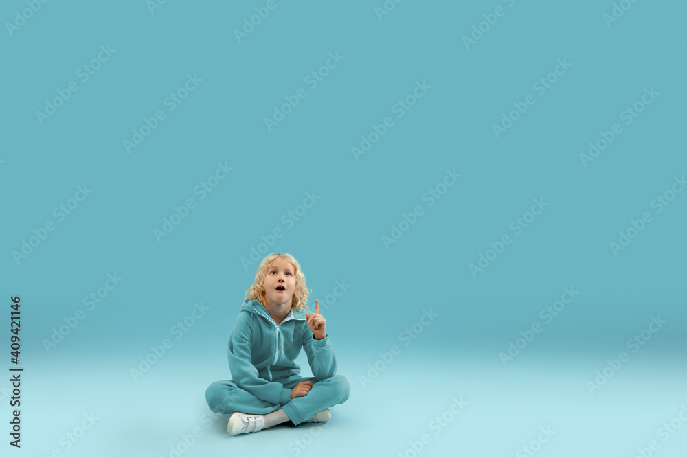 Sitting, pointing. Childhood and dream about big and famous future. Pretty curly boy isolated on blue studio background. Childhood, dreams, imagination, education, facial expression, emotions concept.