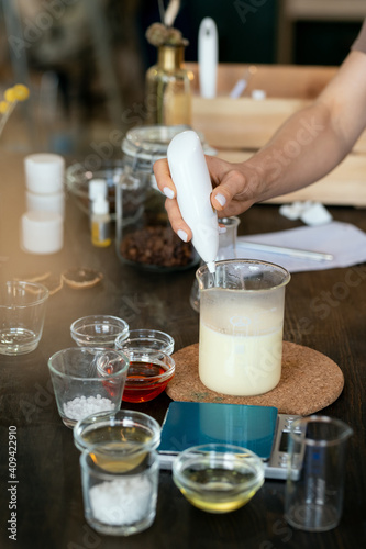 Hand of female putting liquid substance into glassware with melted soap mass