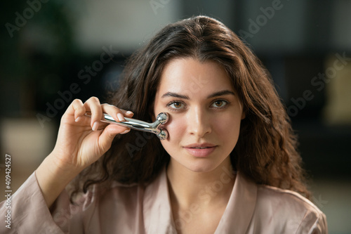 Young woman using metallic massage roller upon her face and looking at you