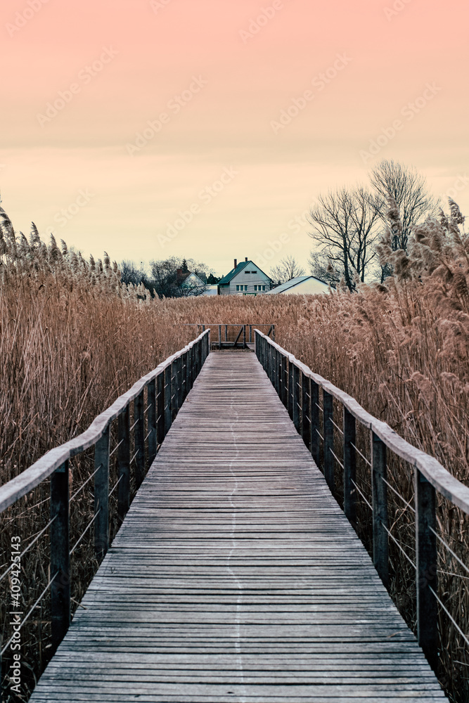 Wooden walkway bridge reaching into a lake. Cute house in the far. Orange and yellow sunset sky. Calm and relaxed mood
