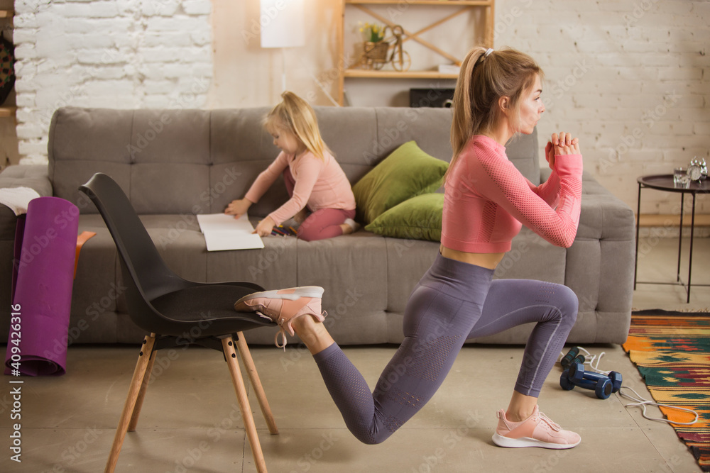 Obraz premium Squats highly concentrated. Young woman exercising fitness, aerobic, yoga at home, sporty lifestyle. Getting active with her child playing, home gym. Healthcare, movement, motherhood concept.