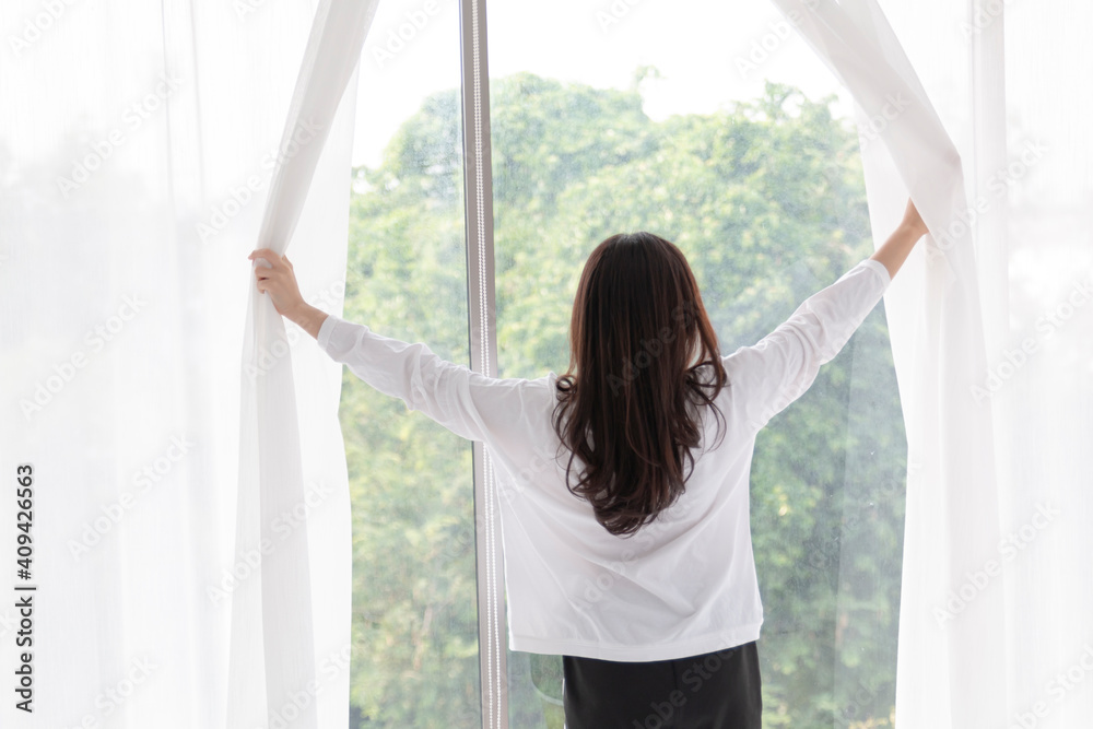 Young woman stand open white curtains at the window in morning after waking up in bedroom. Happy female opening window curtains at home. Woman standing by bedroom window opening curtains. Young woman stand open white curtains at the window in morning after waking up in bedroom. Happy female opening window curtains at home. Woman standing by bedroom window opening curtains.