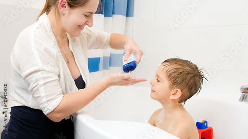 Cute smiling boy taking bath and looking at hi smother washing him with soap and shampoo. Concept of child hygiene and health care at home. Family having time together and playing at home
