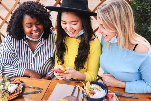 Happy multiracial friends using mobile phone outdoors at brunch restaurant with mask under chins - Focus on asian woman