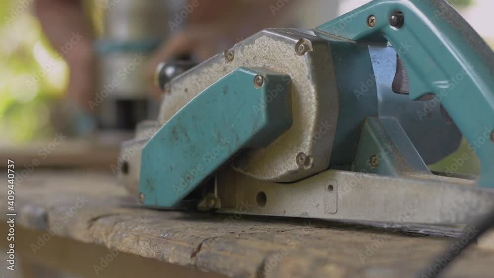 Close up electric planer with old male carpenter working with electric router on wooden slats. Blurred carpenter working on wood craft at workshop to produce construction material or wooden furniture. Close up electric planer with old male carpenter working with electric router on wooden slats. Blurred carpenter working on wood craft at workshop to produce construction material or wooden furniture.