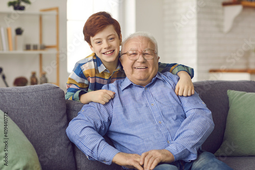 Love and family connection. Family portrait of happy grandfather and little grandson at home in living room. Boy standing behind his grandfather, who is sitting on the couch, hugs his shoulders.