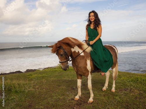 Smiling woman riding horse near the ocean. Outdoor activities. Asia woman wearing long green dress. Traveling concept. Cloudy sky. Copy space. Bali