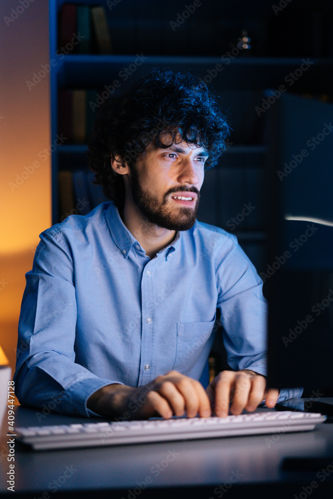 Exhausted young freelancer watching on monitor, working typing on keyboard and drinking hot coffee from cup sitting at desk at home office in dark room. Concept of remote working.