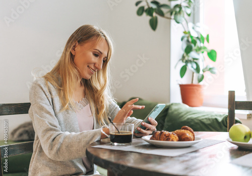 Foto Beautiful blonde young woman having breakfast and using mobile in living room at