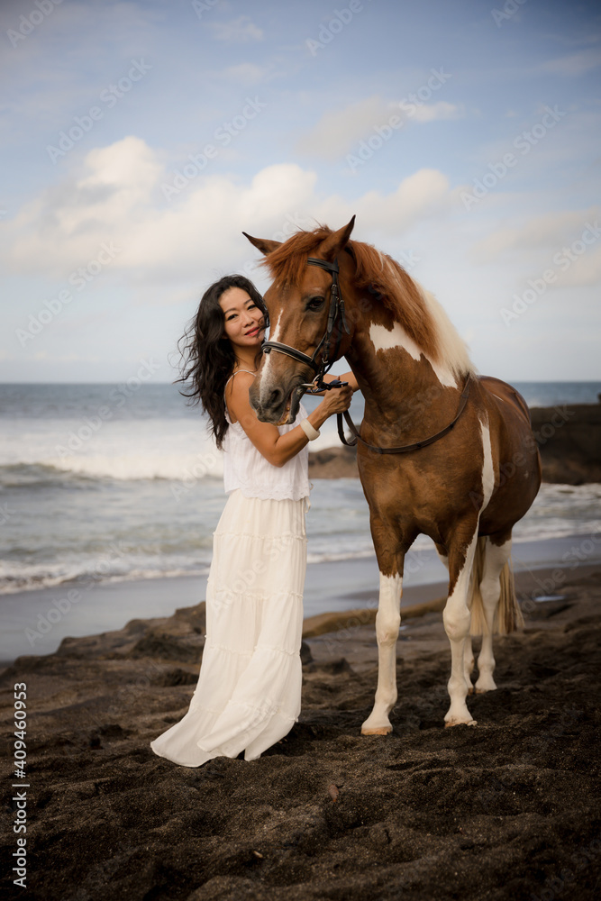 Obraz premium Woman leading horse by its reins. Horse riding on the beach. Love to animals. Asian woman wearing long white dress. Travel concept. Copy space. Bali