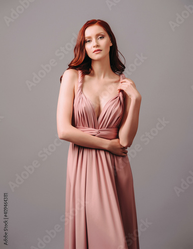 Elegant salmon transformer dress. Beautiful sexy silk evening gown with deep neckline and shoulder straps. Studio touching portrait of young ginger woman. Bridesmaid's fashionable look.