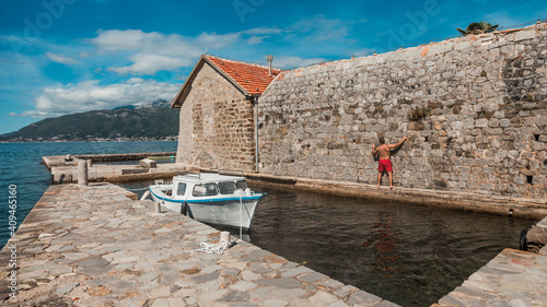 The man is clinging to a stone wall of a medieval church in Montenegro. A small private boat at the pier and beautiful mountains in the background. Island Gospa od Milosti in Montenegro photo