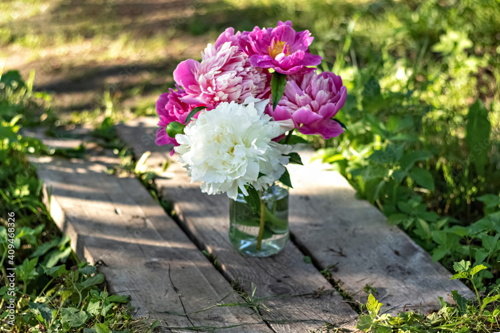 A bouquet of large white and pink peonies in a glass jar on a wooden bridge in the garden. Bloom