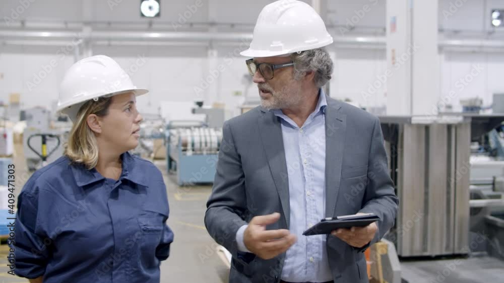 Senior factory boss with tablet commenting machinery work process to female engineer. Colleagues in hardhats walking on plant floor and discussing equipment. Industrial occupation concept Senior factory boss with tablet commenting machinery work process to female engineer. Colleagues in hardhats walking on plant floor and discussing equipment. Industrial occupation concept