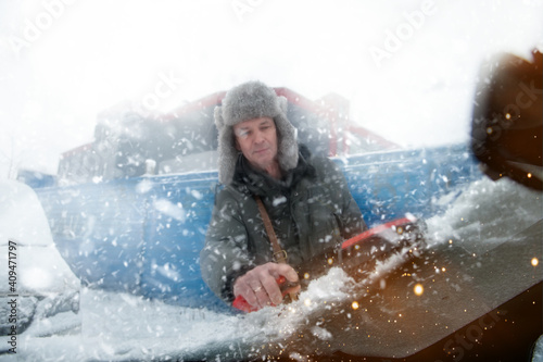 Man in a fur hat cleans snow from the roof of a car outdoors on a winter day
