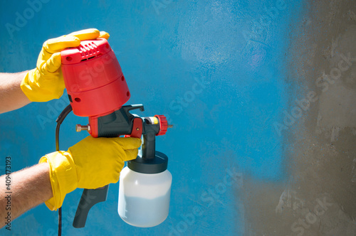A worker's hands in yellow gloves holds a hand-held spray gun with an overhead compressor. Painting the wall of the house in blue. Household renovation. Copy space. Selective focus photo