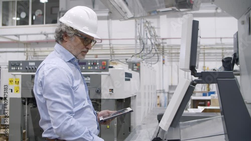 Senior engineer testing industrial machine on plant floor, pressing buttons at control board, monitoring process, using tablet. Production process or machinery concept
