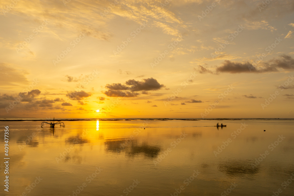A moored fishing boat and kayak silhouetted against a reflected setting sun and golden sky from Nusa Dua beach in Bali.