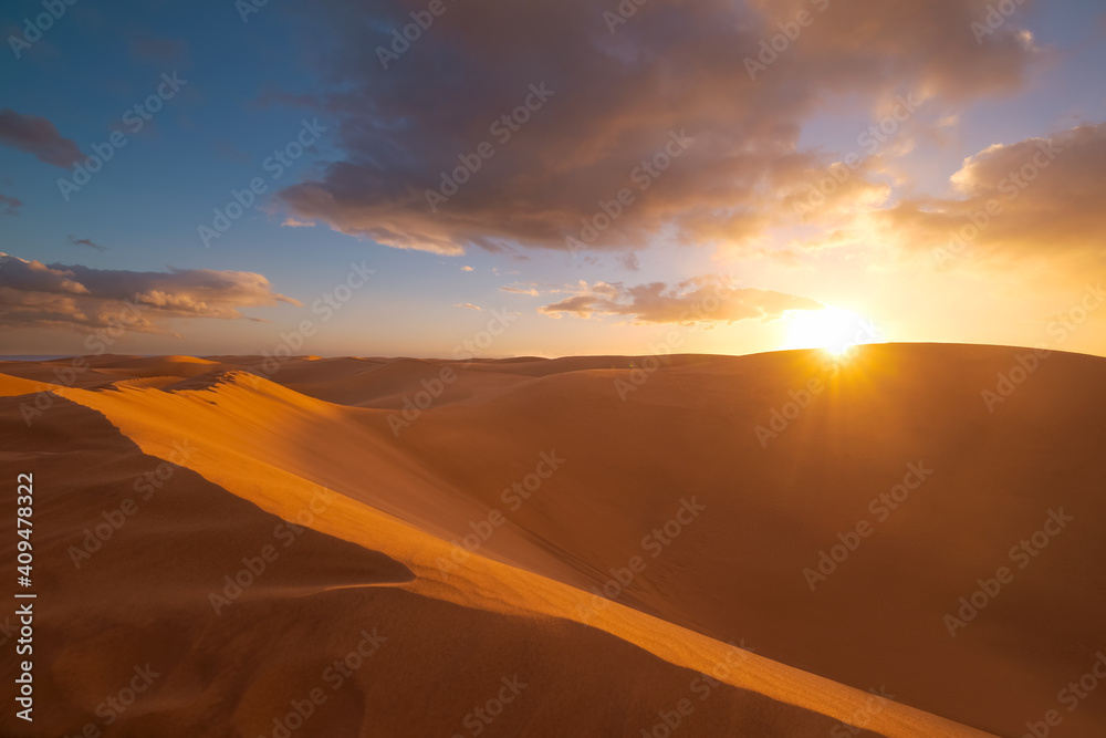 Golden sand dunes in desert in Maspalomas. Sunset in the desert, sun and sunrays, beautiful dramatic clouds and blue sky. Gran Canaria, Canary islands, Spain