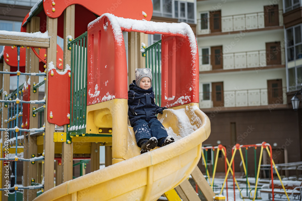 Little cute Caucasian boy wearing knit scandinavian hat and winter overall sliding down a plastic slide  at the playground in winter.