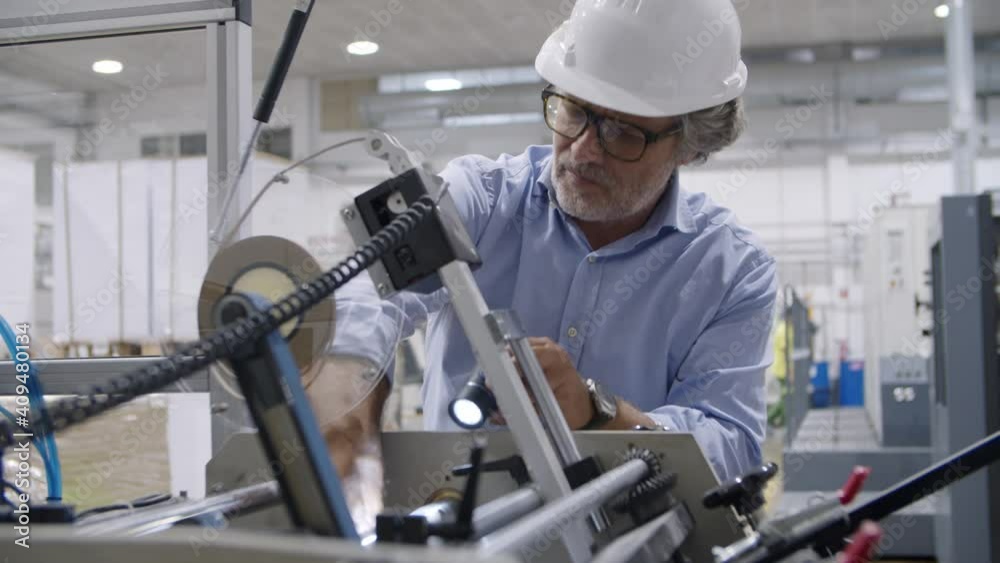 Focused mature factory technician wearing hardhat, using torch, examining and repairing industrial machine on plant floor. Production or machinery concept