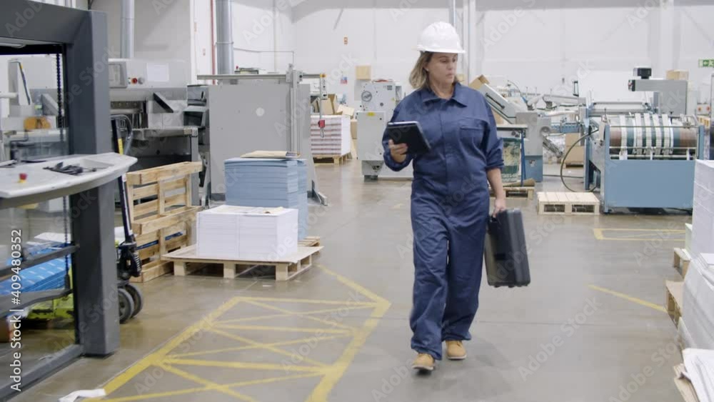 Middle aged female factory engineer with toolkit box walking on plant floor, looking around, holding and using tablet. Dolly shot. Industry and production concept Middle aged female factory engineer with toolkit box walking on plant floor, looking around, holding and using tablet. Dolly shot. Industry and production concept