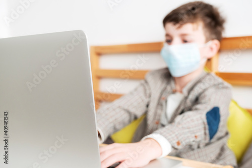 Boy medical mask is watching school lesson laptop, sitting bed on pillows. Quarantine in world Coronavirus COVID-19 virus. Epidemic, pneumonia, hygiene. Pandemic. Distance learning. Online education