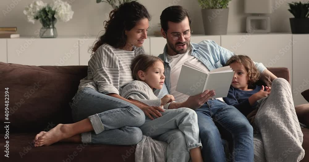 Full family gathered in cozy living room, couple and kids resting on comfy couch reading fairy tale to little curious attentive son and daughter. Development education of children, fun, hobby concept Full family gathered in cozy living room, couple and kids resting on comfy couch reading fairy tale to little curious attentive son and daughter. Development education of children, fun, hobby concept