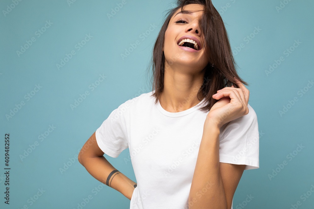 Fototapeta premium Photo of young beautiful happy smiling brunette woman wearing trendy white t-shirt with empty space for mock up. Sexy carefree female person posing isolated near blue wall in studio with free space