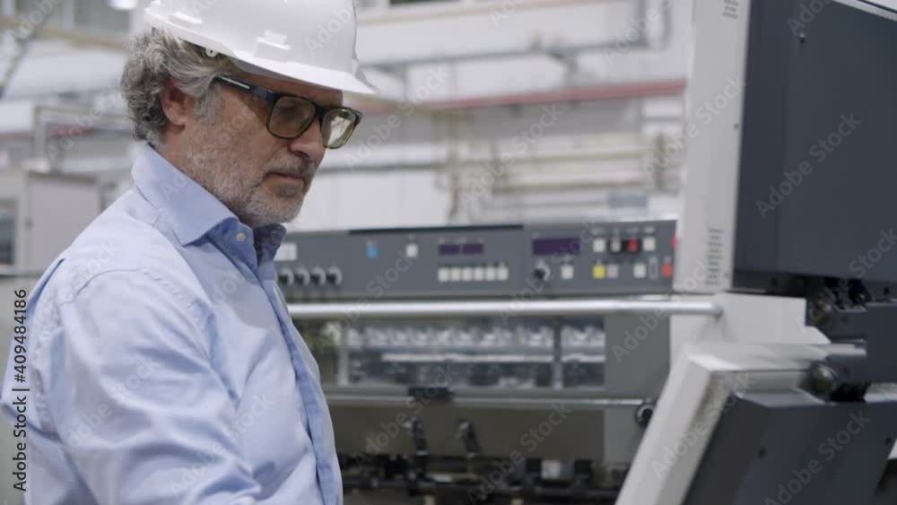 Older engineer checking and operating industrial machine on plant floor, pressing buttons at control board, monitoring process, using tablet. Production process or machinery concept Older engineer checking and operating industrial machine on plant floor, pressing buttons at control board, monitoring process, using tablet. Production process or machinery concept