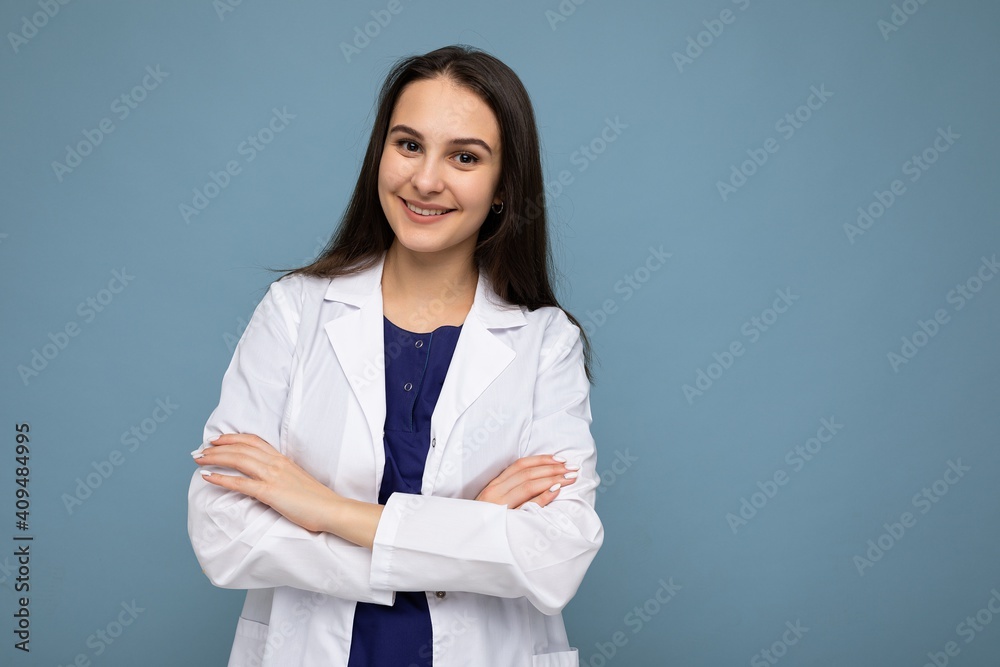Photo portrait of young pretty beautiful positive smiling brunette woman with sincere emotions wearing white medical coat isolated over blue background with copy space and holding crossed arms Photo portrait of young pretty beautiful positive smiling brunette woman with sincere emotions wearing white medical coat isolated over blue background with copy space and holding crossed arms