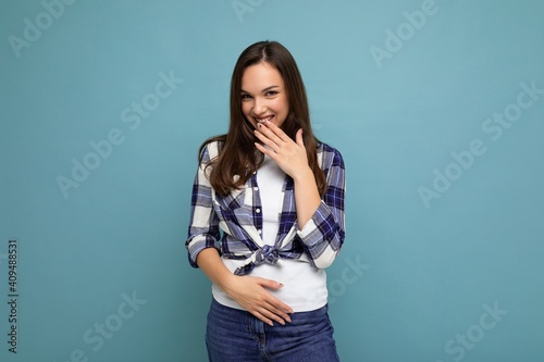 Young positive happy beautiful winsom brunette woman with sincere emotions wearing check shirt poising isolated over blue background with copy space and having fun