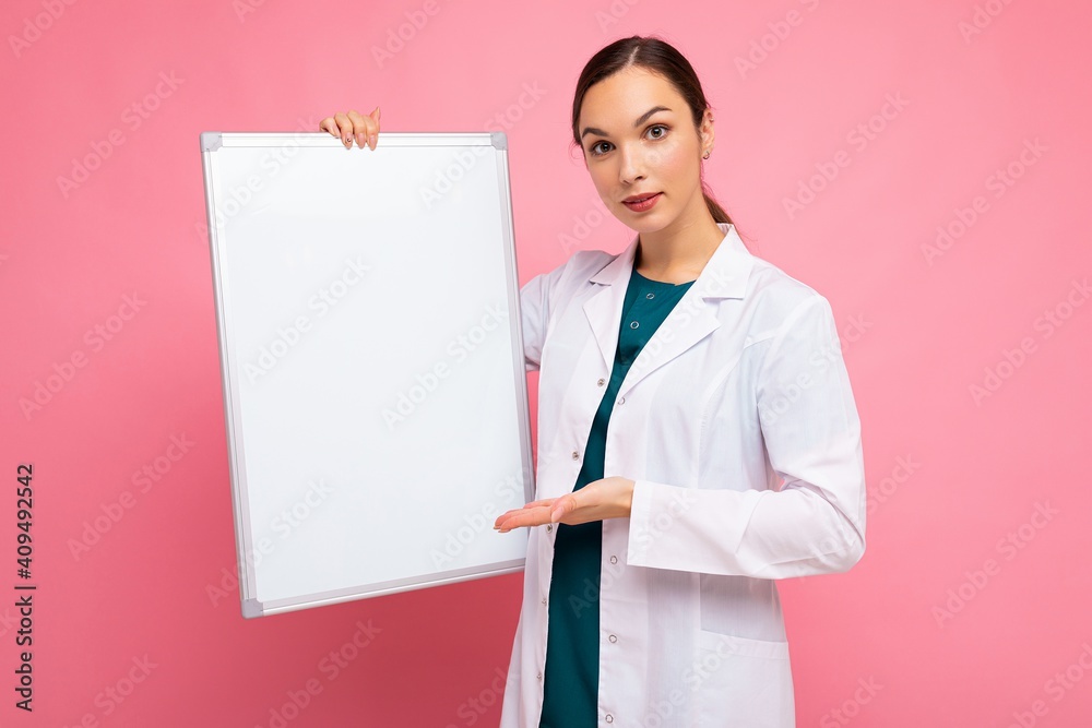 Portrait of attractive pretty young positive brunette woman wearing medical white coat holding white magnetic board for mock up isolated over pink background with copy space Portrait of attractive pretty young positive brunette woman wearing medical white coat holding white magnetic board for mock up isolated over pink background with copy space