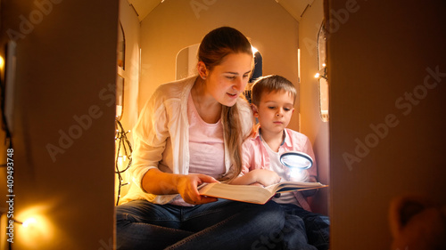 Portrait of smiling young mother with little boy reading book at night while playing in toy house. Concept of child education and family having time together at night