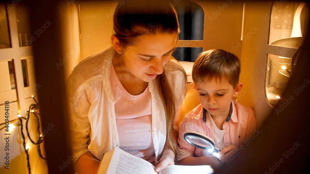 Portrait of cute boy with young mohter reading big book in tent or toy house before going to sleep. Concept of child education and family having time together at night