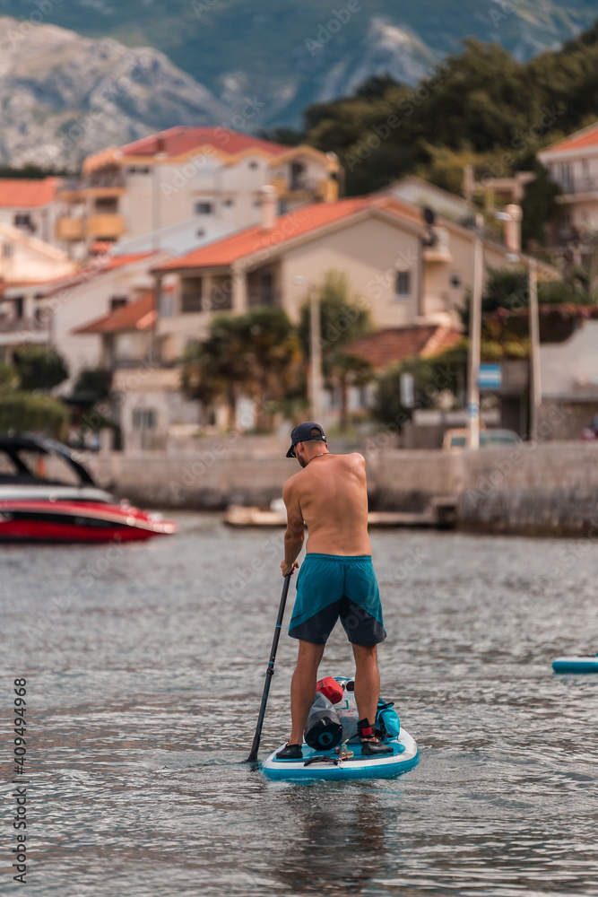 A young athletic man stands on a SUP stand up paddle board in Kotor Bay overlooking a beautiful village in Montenegro