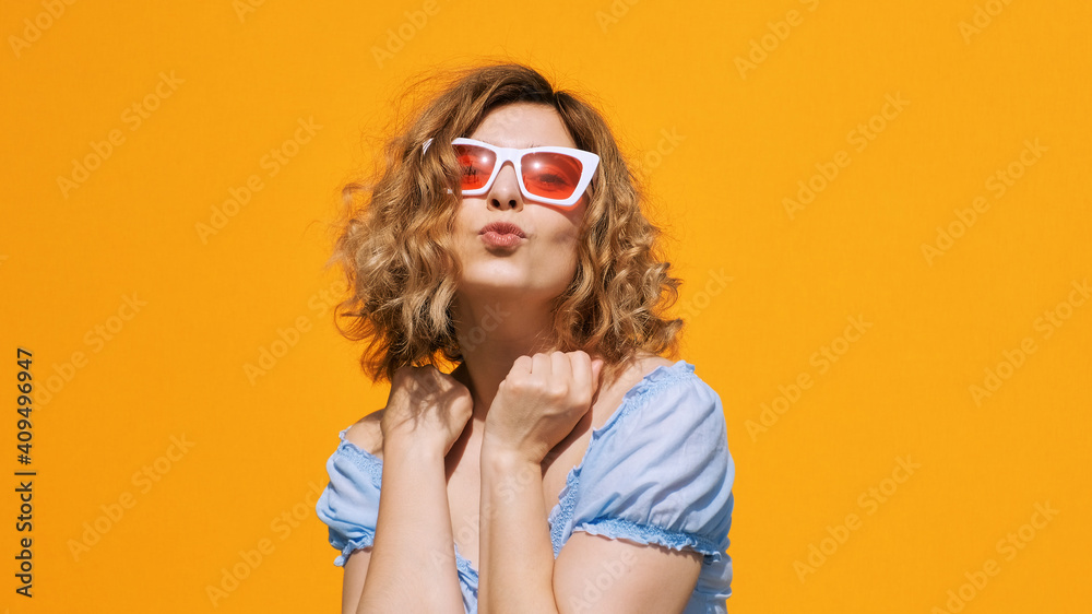 Portrait of happy woman in stylish white sunglasses red glasses blonde with curly hair, smiling with kisses looking on camera yellow background in summer. Emotions. Positive girl. Freedom. Lifestyle