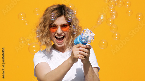 Portrait of happy woman in stylish sunglasses with soap bubbles smiling releases toys from gun with pleasure on yellow background in summer. Emotions, joy. Positive flirty girl. Childhood