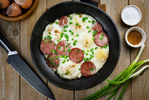 Fried eggs with sausage and green onions in a frying pan on a wooden table