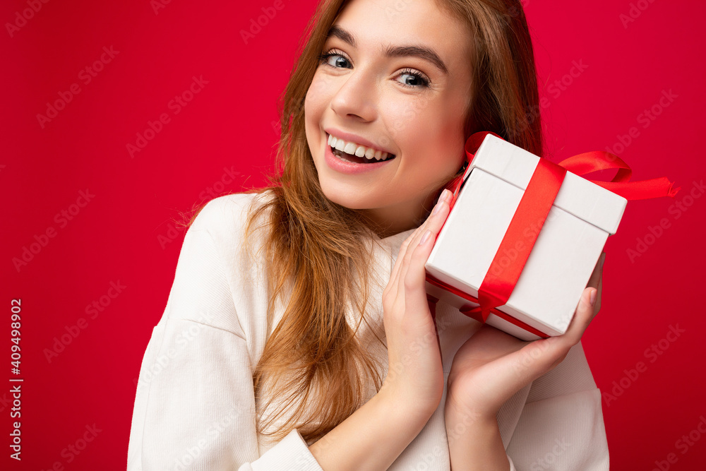closeup portrait photo shot of attractive delightful smiling blonde young woman isolated over red background wall wearing casual white hoodie holding white gift box with red ribbon and looking at closeup portrait photo shot of attractive delightful smiling blonde young woman isolated over red background wall wearing casual white hoodie holding white gift box with red ribbon and looking at