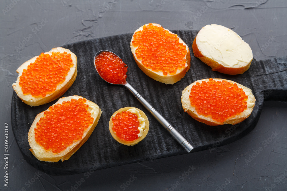 Canapes with butter and red caviar next to a spoon with caviar on a wooden board on a concrete background.