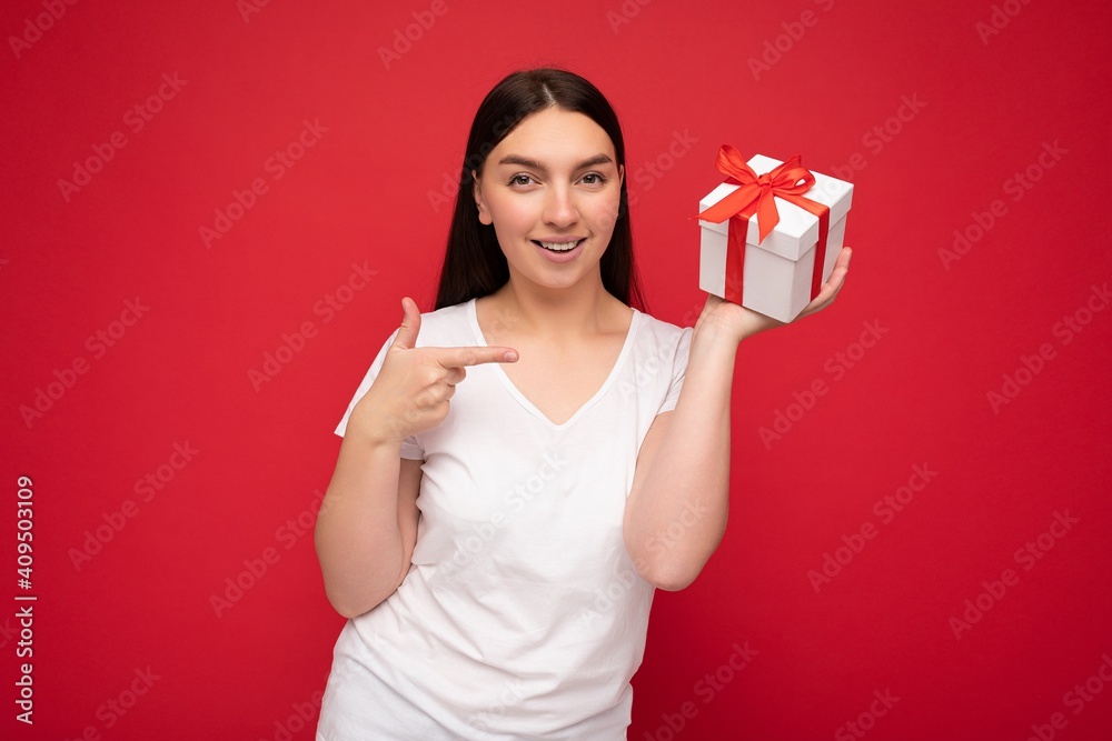 Photo of beautiful positive smiling young brunette woman isolated over red background wall wearing white casual t-shirt for mockup holding white gift box with red ribbon and looking at camera and Photo of beautiful positive smiling young brunette woman isolated over red background wall wearing white casual t-shirt for mockup holding white gift box with red ribbon and looking at camera and