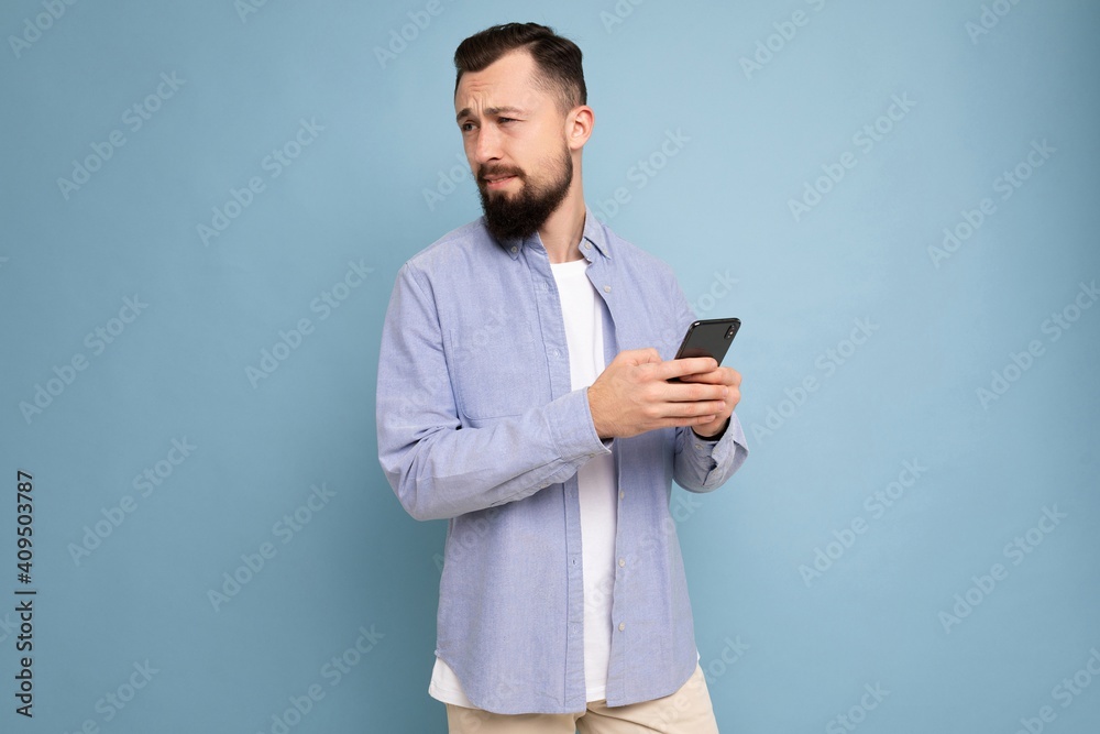 Thoughtful handsome young man isolated over background wall wearing everyday clothes holding and using mobile phone writing sms looking to the side and thinking Thoughtful handsome young man isolated over background wall wearing everyday clothes holding and using mobile phone writing sms looking to the side and thinking