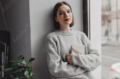 Brown-eyed dark blond girl with red lipstick dressed in gray sweater firmly holding book and posing leaning on wall